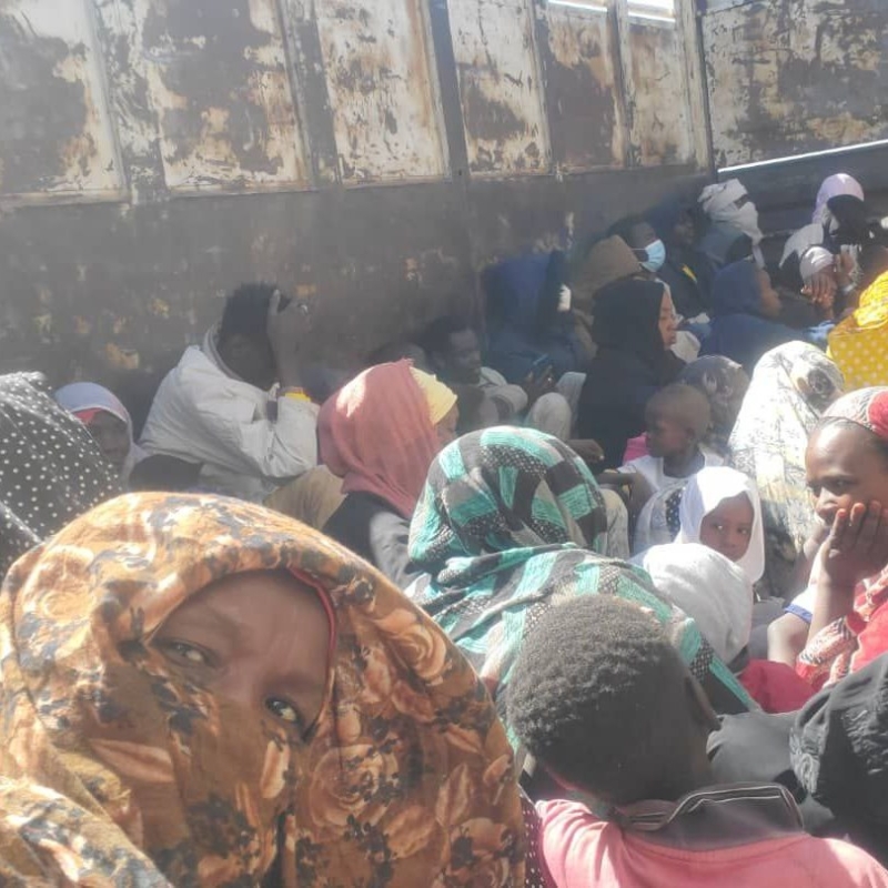 Fatima, in a floral headscarf, crouches with a group of women and children inside a rusty metal container, seeking shelter from gunfire. Some cover their ears or faces. Expressions show fear and tension under harsh daylight.