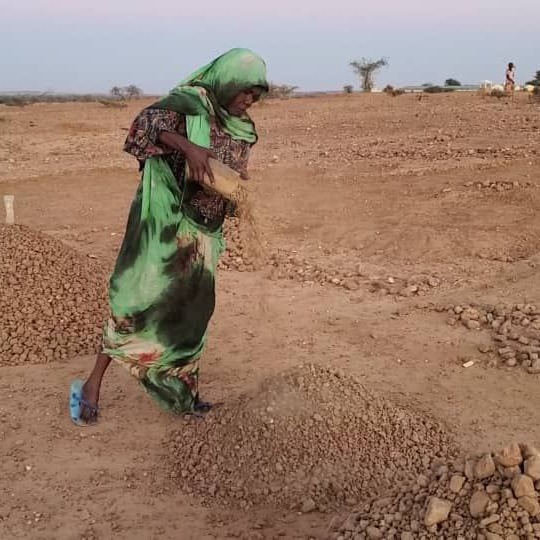 Fatima wears a green headscarf and a patterned dress while extracting gravel into a pile in dry, rocky terrain.