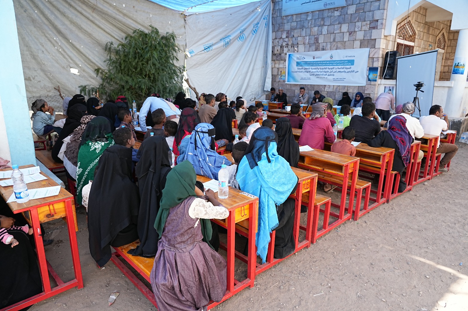 A group of Yemeni children and parents sit at red wooden school desks under a tent and beside a stone building, attending an outdoor workshop. Some wear headscarves, water bottles and papers are on desks, and a banner with Arabic text hangs in the background.