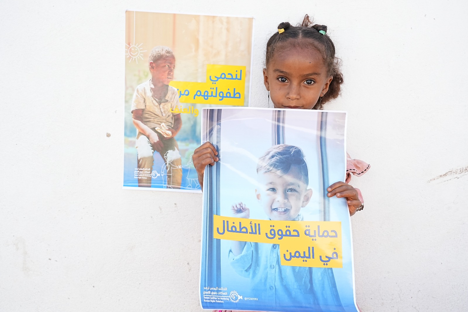 A young brown girl with curly hair in braids holds a poster showing a smiling child, with Arabic text reading “Protecting Children’s Rights in Yemen.” Behind her, another poster shows a boy and Arabic text. She looks directly at the camera against a plain white wall.