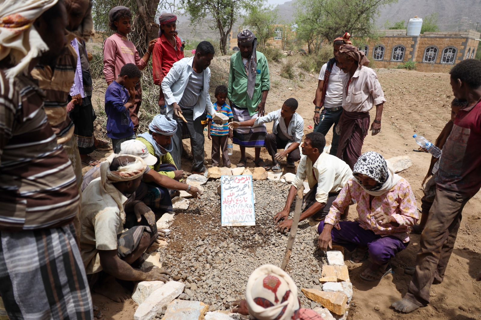 A group of men and boys are gathered outdoors in a dry, rocky terrain for a burial ceremony. Several men are crouched around a freshly dug grave lined with stones, while others stand nearby observing the solemn event. A makeshift white sign placed on the gravel mound identifies the deceased as Ahmed Saif Abdullah Al-Wafah. The sign also includes handwritten dates, marking his birth on 25/8/2020 and his death on 29/11/2025, indicating the burial of a young child. The attendees wear traditional clothing and turbans, set against a backdrop of sparse trees and a stone building.