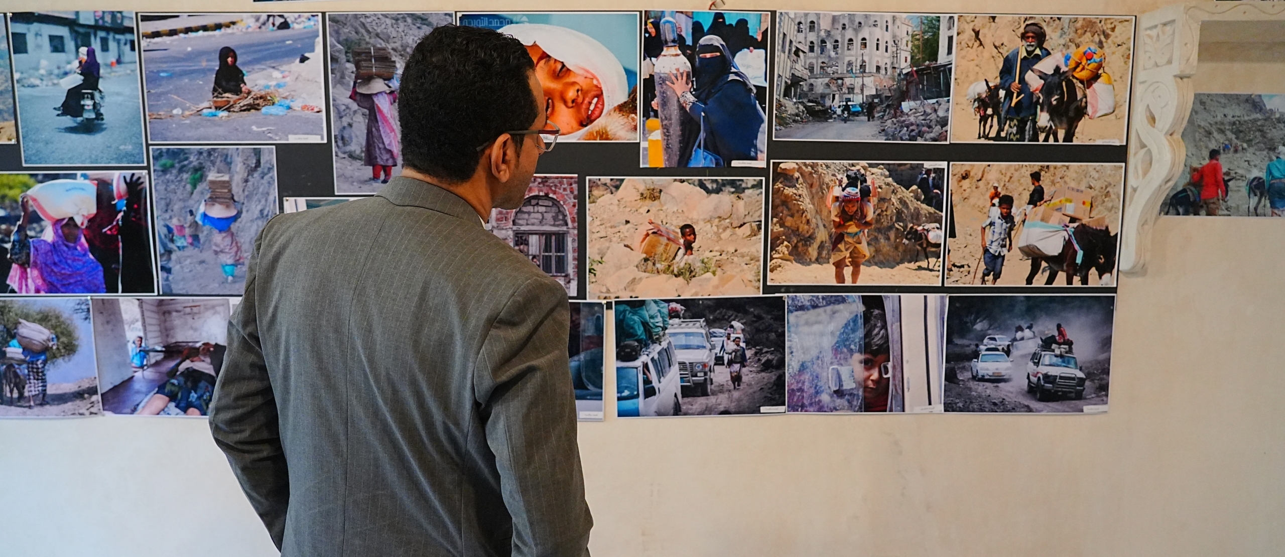 A man in a suit stands facing a wall display of documentary photographs showing scenes of conflict, displacement, and daily life, viewing the images as part of a memory museum exhibition.