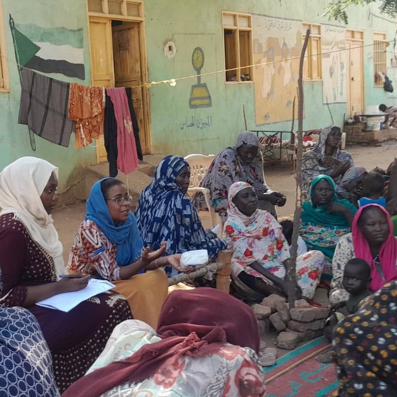 Women in colorful headscarves sit and stand outside a painted light green building with laundry hanging on a line, “Equality for all” is written in Arabic alongside a mural. Some women are holding papers and all are listening to one of them speak.