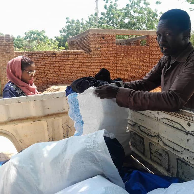 A man in a brown shirt loads large white sacks into the back of a pickup truck, while a woman in a pink headscarf stands nearby. Behind them stands brick walls and trees under bright daylight.