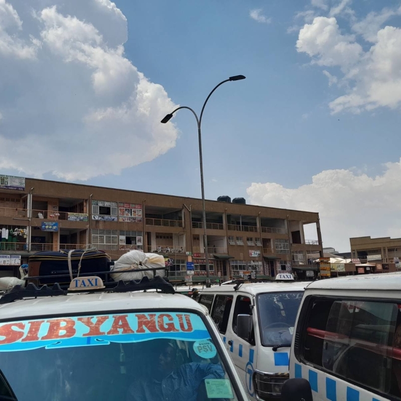 White taxis with “SIBYANGU” and “TAXI” signs parked in front of a multi-story building under a partly cloudy sky. Luggage is strapped to roofs and people are visible inside and around the vehicles. A building in the back is a mix of residential and commercial units.