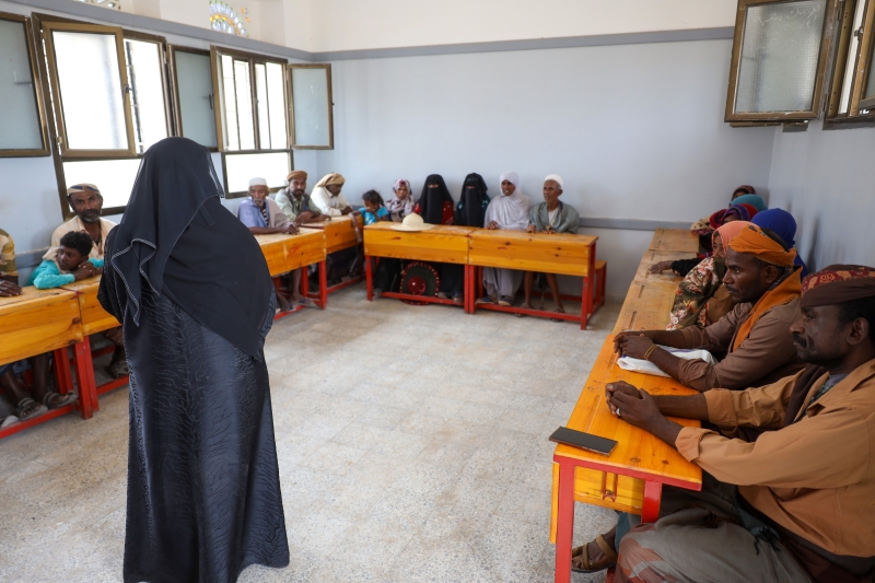 A veiled woman stands at the front of a simple classroom, addressing a group of men and women seated on wooden benches arranged in a circle, as they listen and participate in a community discussion.