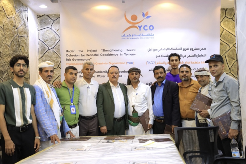 A group of participants stand together behind a table during a Social Cohesion and water conflict resolution workshop hosted by the Youth Creativity Organization, posing for a group photo in front of the event banner.