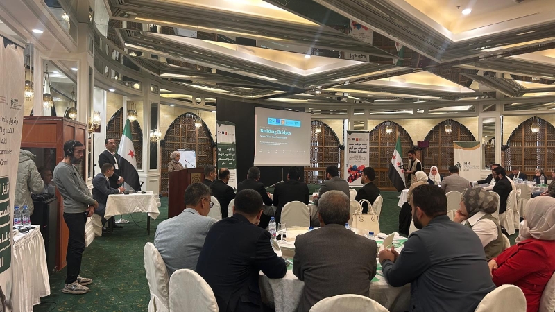 A conference session in progress with a speaker at a podium addressing an audience seated at tables. A projection screen reads "Building Bridges: Seven Cities, One Conversation." Syrian flags are displayed on either side of the stage. Banners include logos for "IHR International Humanitarian Relief" and Arabic text. Attendees are focused on the presentation, and the room features arched windows and ornate decor.