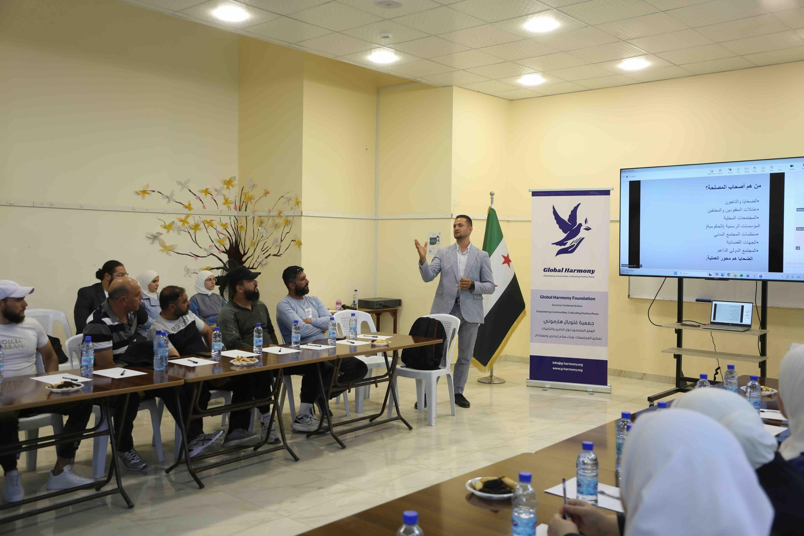 One of DTI’s Senior Program Managers, Feras Hamdouni, wears a light grey suit while standing at the front of a conference room, gesturing with his right hand as he presents to an audience. Behind him is a Syrian flag and a banner for the "Global Harmony Foundation." A large screen to his right displays a presentation slide with Arabic text. The audience, a mix of men and women, is seated at long tables arranged in a U-shape, with water bottles, notepads, and small plates of snacks in front of them. A decorative tree mural is visible on the wall in the background.