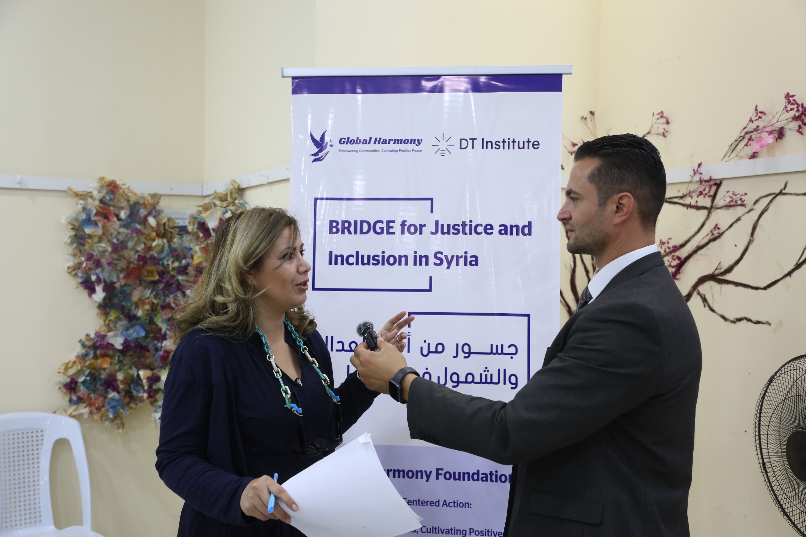 A woman with blonde hair, wearing a dark blue top and a turquoise necklace, is being interviewed by a man in a dark suit. The man holds a small microphone towards her as she speaks, holding a piece of paper and a pen. They stand in front of a banner for "Global Harmony" and "DT Institute" that reads "BRIDGE for Justice and Inclusion in Syria" in both English and Arabic. Colorful, abstract wall art is visible behind them.