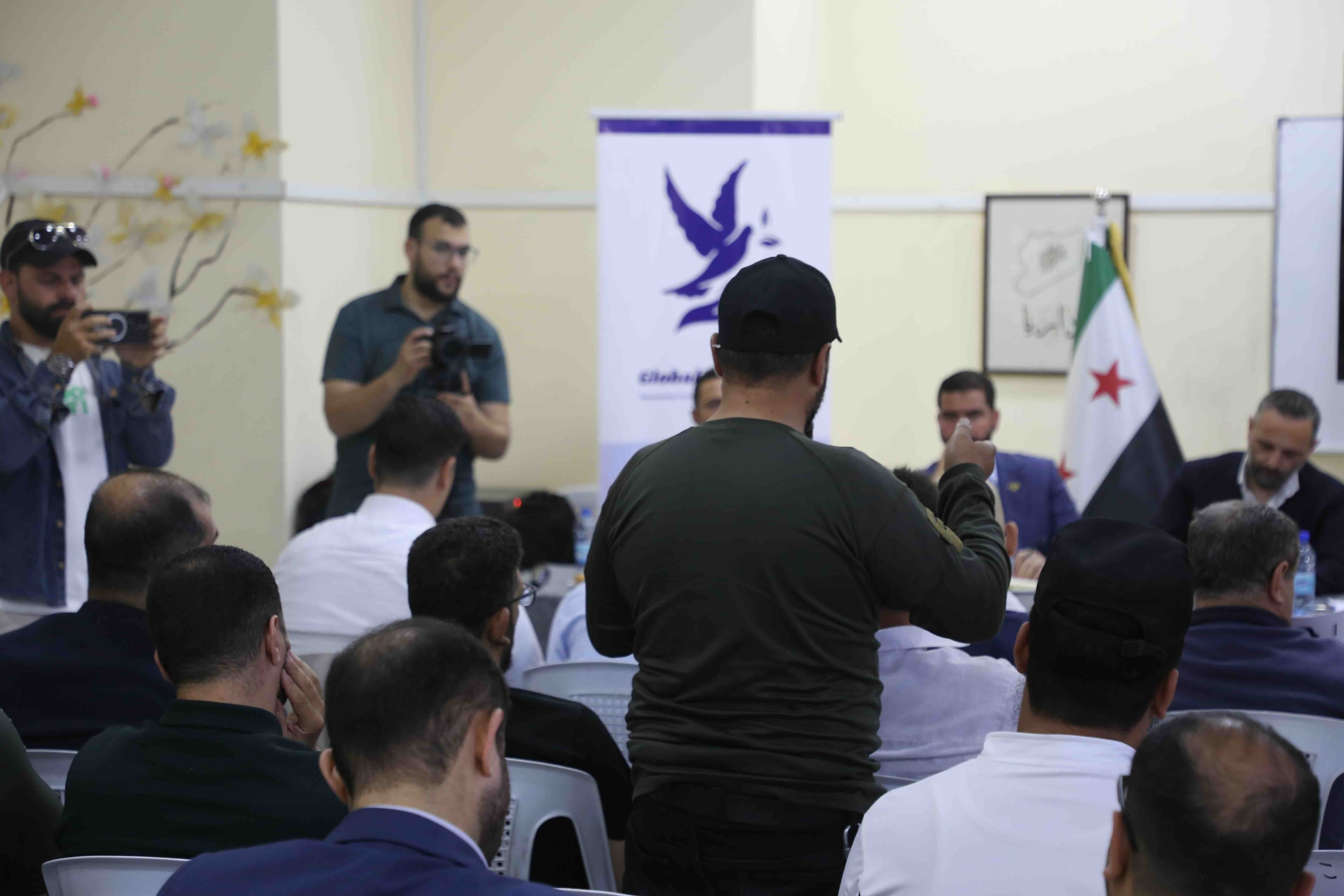 A view from behind an audience of men seated in a conference room. In the foreground, a man in a black t-shirt and baseball cap stands with his back to the camera, raising his hand as if to ask a question. In the background, two men stand holding cameras, documenting the event. A Syrian flag and a banner with a purple dove logo are visible behind a panel of seated men.