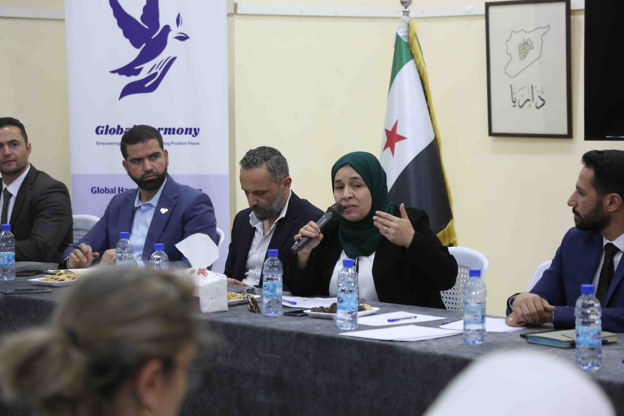 A panel of five people, four men and one woman, are seated at a long table covered with a dark cloth. The woman, wearing a green hijab and a black blazer, is speaking into a microphone and gesturing with her left hand. The men on the panel are listening attentively. On the table are water bottles, papers, pens, and small plates of cookies. Behind them, a "Global Harmony" banner and a Syrian flag are displayed. A framed piece of art showing a map of Syria with a bunch of grapes is on the wall.