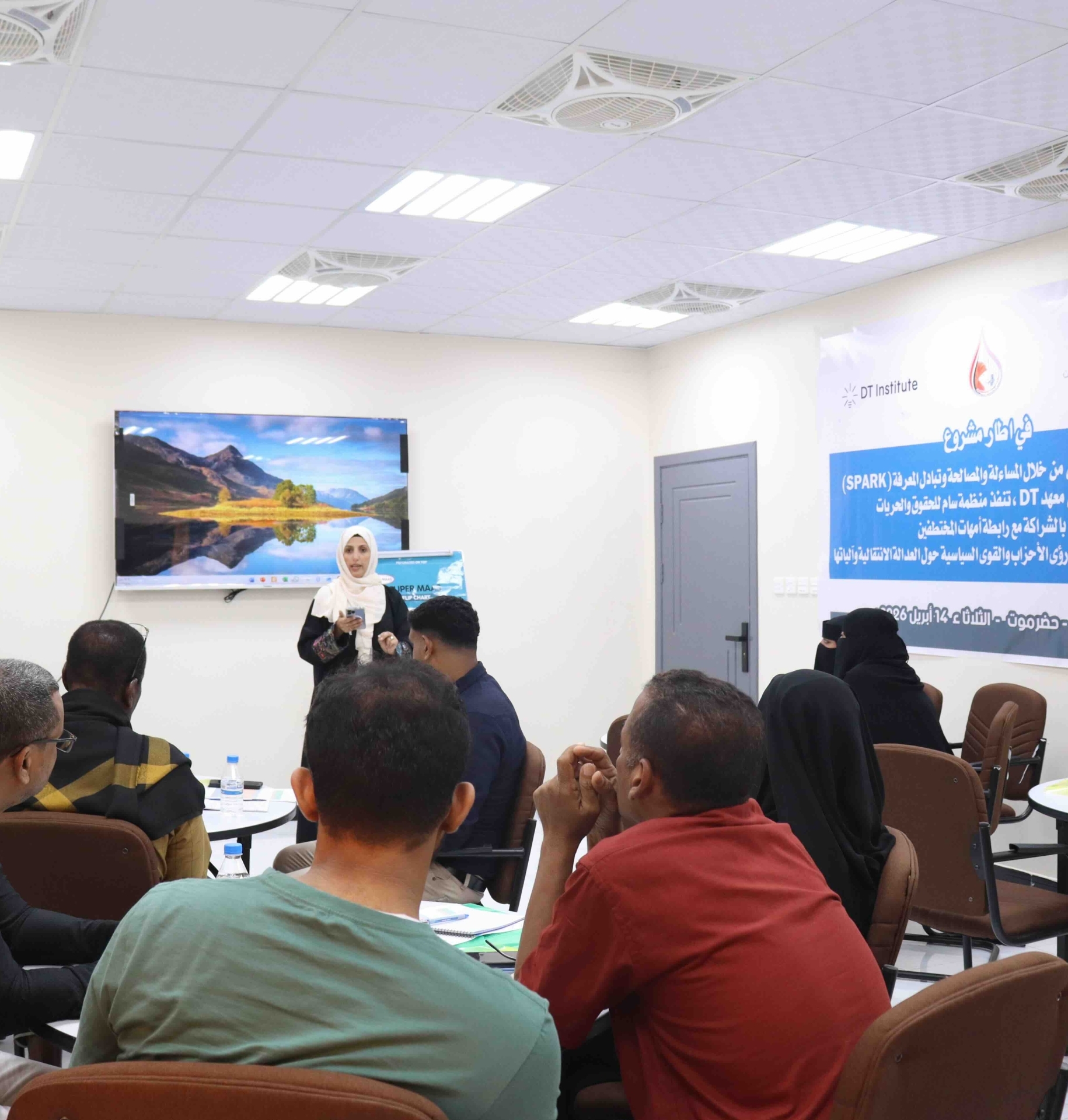 Inside a well-lit conference room, a woman wearing a white hijab and black attire stands at the front, addressing a seated audience. Behind her, a large screen displays a scenic landscape of mountains and water, while a banner to the right features logos for "DT Institute" and "SAM" alongside Arabic text regarding the "SPARK" project and support for peace in Yemen. The audience, comprising men in casual clothing and women in black abayas, is seated at round tables with water bottles and notebooks, listening attentively to the presentation.