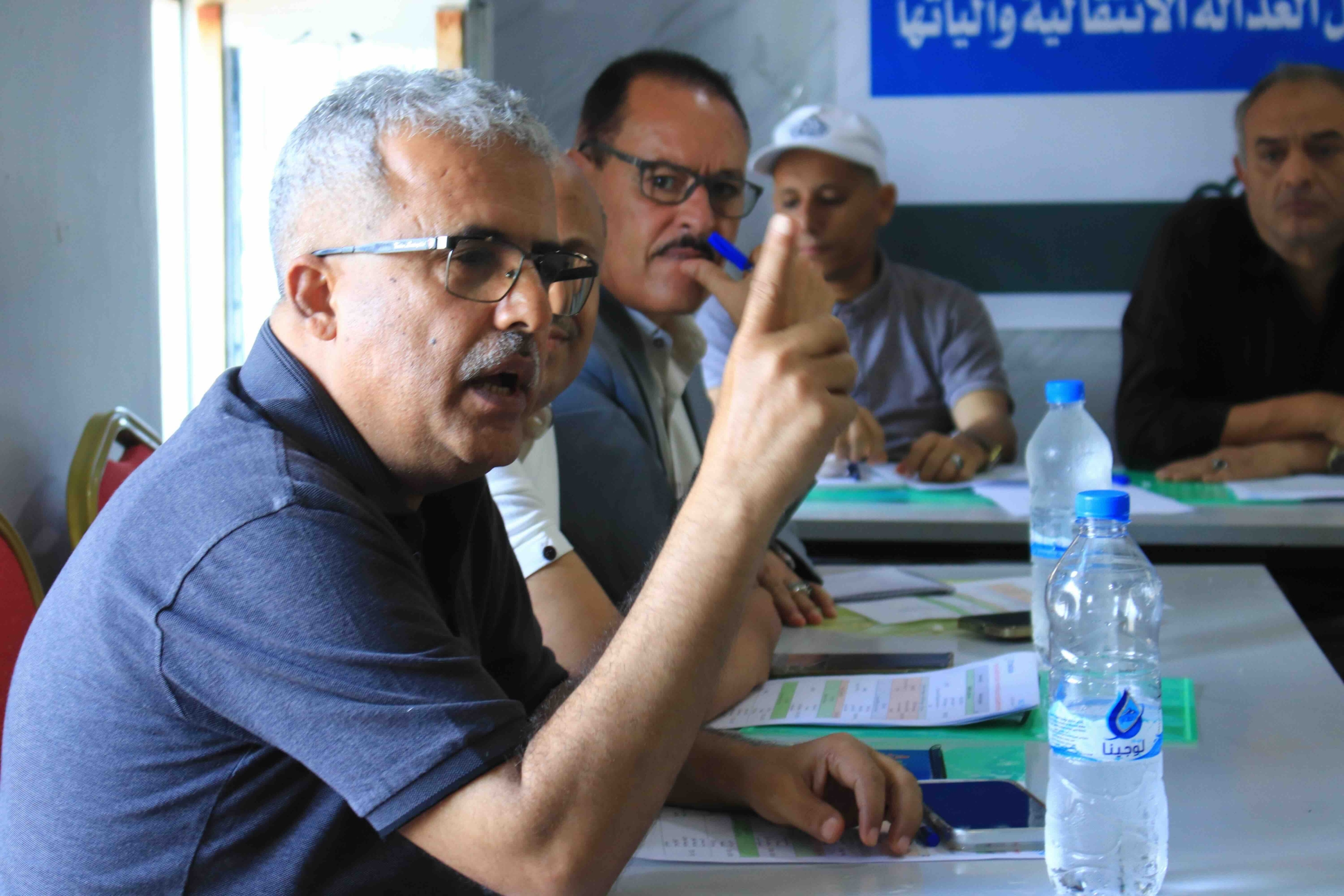 A close-up shot captures a man with graying hair and glasses speaking passionately during a meeting. He is wearing a blue polo shirt and gesturing with his right index finger raised to emphasize a point. Seated at a table with papers and water bottles, he is surrounded by other men who are listening intently; one man in the background holds a blue pen to his lips in thought. A blue banner with white Arabic text is partially visible on the wall behind them, indicating a formal discussion or workshop setting.