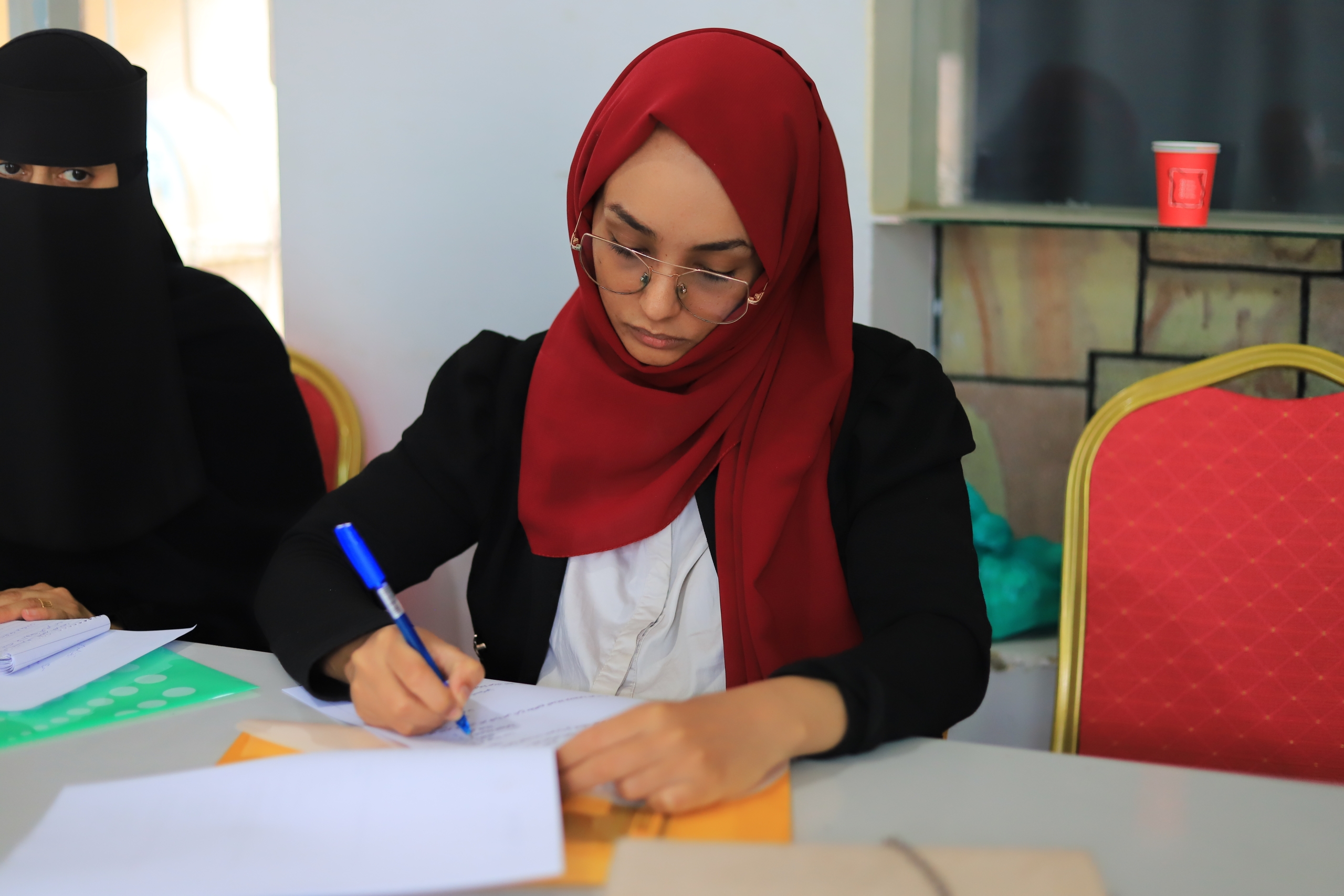 A young woman wearing a red hijab, glasses, and a black blazer is seated at a table, focused on writing on a document with a blue pen. To her left, another woman wearing a black niqab is partially visible, also seated at the table with papers. The meeting room shows a red and gold chair in the foreground and a red cup on a shelf in the background.