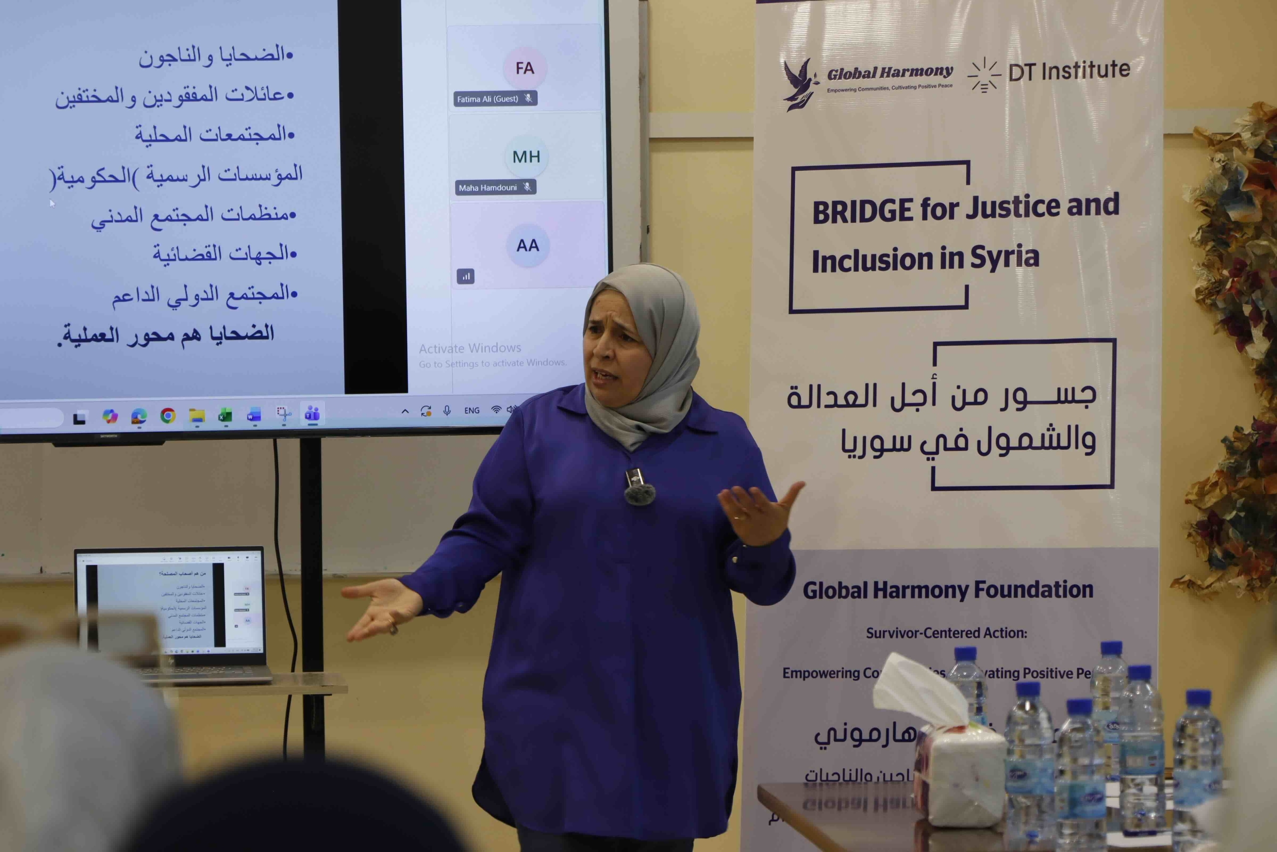 A woman wearing a grey hijab and a purple shirt stands in front of a large screen, gesturing with both hands as she speaks. The screen displays a presentation slide with Arabic text and a video call interface. To her right is a banner for "Global Harmony" and "DT Institute" with the title "BRIDGE for Justice and Inclusion in Syria" in English and Arabic. A laptop on a small table to her left mirrors the presentation slide, and several water bottles and a tissue box are on a table in the foreground.