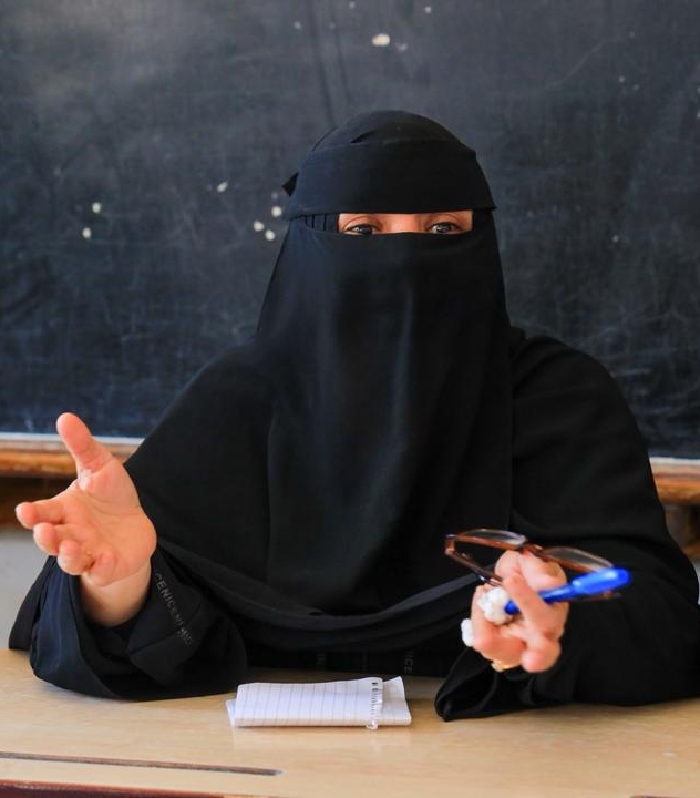 A woman wearing a black niqab and abaya is seated at a wooden desk in front of a dark chalkboard, actively participating in a discussion. She gestures with her right hand while holding a pair of glasses and a blue pen in her left. A small spiral notebook and a smartphone rest on the desk before her. To her right, the arm of another person in a green sleeve is visible.