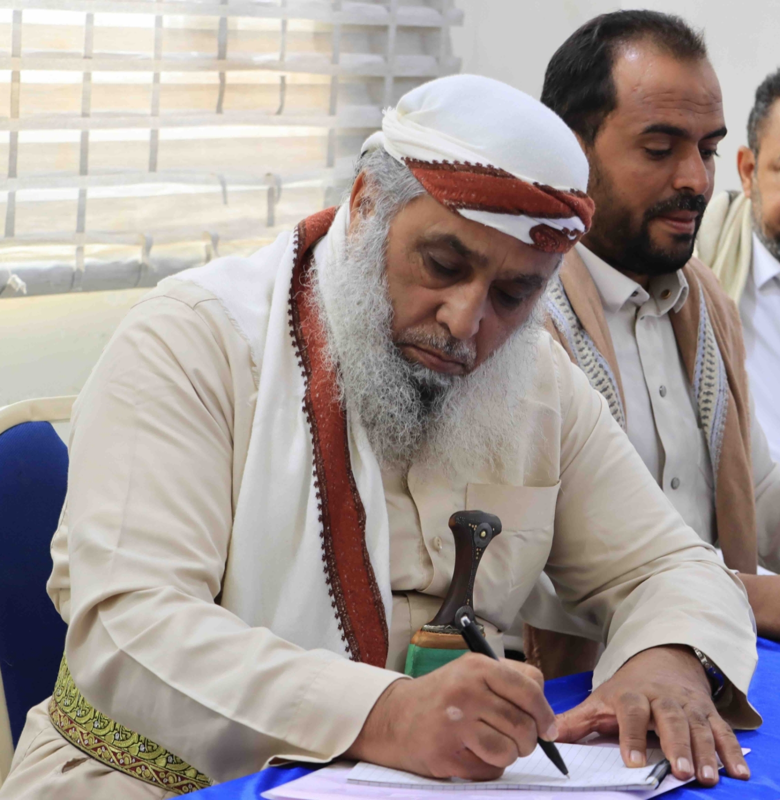 An elderly man with a long white beard, wearing a traditional white thobe, an embroidered belt, and a white turban with a red band, is seated at a table covered with a blue cloth. He is focused on signing a document with a black pen. A ceremonial dagger (jambiya) is tucked into his belt. Two other men sit beside him, observing the signing closely. Blinds cover the windows in the background.