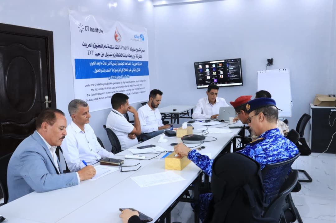 A group of men are seated around a long white conference table in a bright, modern meeting room. On the wall behind them is a large banner with Arabic and English text for the "DT Institute" and the "SPARK Project," indicating a panel discussion on "Community Reconciliation" held in July 2025. Some attendees are in civilian suits and shirts, while others wear military or police uniforms, including one man in a blue camouflage uniform and another in a red beret. A laptop on the table displays a video conference grid, suggesting a hybrid meeting format.