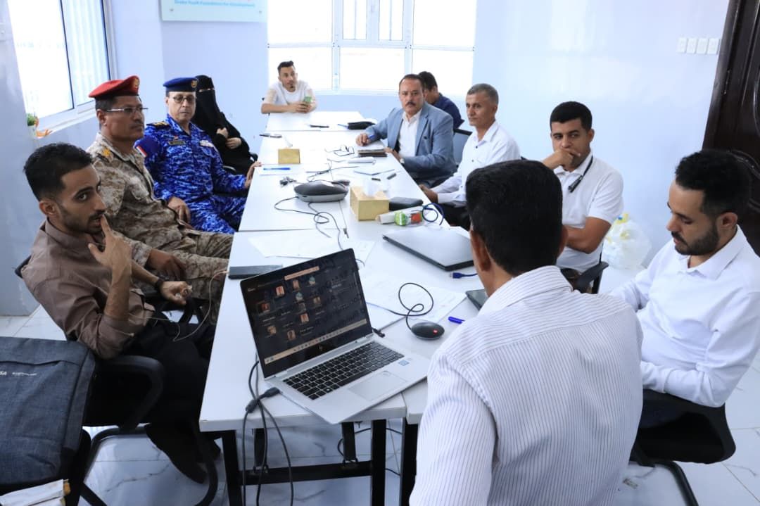 A wide angle of the same meeting shows the full length of the table with diverse attendees engaged in discussion. In the foreground, a man in a striped shirt faces away from the camera, looking toward a laptop showing a video call interface. To the left, men in military fatigues and a red beret listen intently. At the far end of the room, a woman in a black niqab is seated near a window, participating in the gathering. The scene depicts a collaborative session involving civil and security stakeholders.